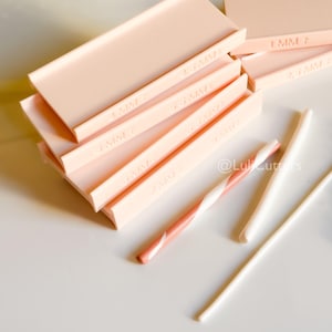 Könnte beinhalten: Eine Sammlung hellrosa Polymer-Tonwerkzeuge, darunter rechteckige Ausstecher mit Millimeter-Maßen. Ein Werkzeug aus Ton mit Zuckerstangenmuster und zwei weiße Tonwerkzeuge sind ebenfalls vorhanden. Die Werkzeuge sind auf einer weißen Oberfläche angeordnet.