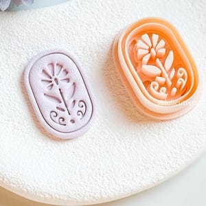 May include: Two floral-shaped cookie cutters. One is light purple, the other is peach. Both are rectangular with rounded corners and feature a detailed flower design. The cutters are on a white, textured surface.