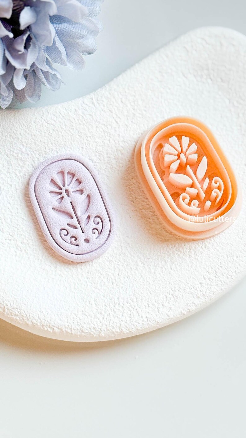 May include: Two floral-shaped cookie cutters. One is light purple, the other is peach. Both are rectangular with rounded corners and feature a detailed flower design. The cutters are on a white, textured surface.