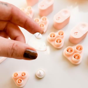 May include: A collection of light pink and white ear piercing tools. One is held by a hand, showing a clear, triangular earring. Other tools are in the background, including pink, oval-shaped pieces with numbers and triangular pieces with three holes.