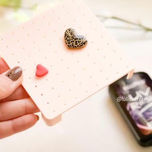 Może przedstawiać: Różowy plastikowy stojak do suszenia z otworami do suszenia kolczyków z polimerowej gliny. Na stojaku znajdują się dwa kolczyki w kształcie serca, jeden z nadrukiem lamparta, a drugi jednokolorowy czerwony.