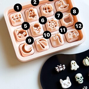 May include: A collection of Halloween-themed cookie cutters in a light pink tray. The cutters include shapes like a spider, ghost, pumpkin, witch's hat, cat, skull, spiderweb, cauldron, crescent moon, candy cane, and a video game controller.