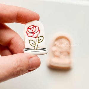 May include: A white cookie cutter in the shape of a glass dome with a red rose inside. The rose has green leaves and stem. The cookie cutter is held in a hand. Another cookie cutter is in the background.