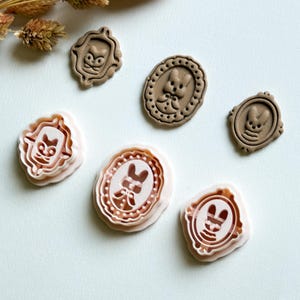 May include: A collection of brown and pink stampers featuring whimsical bunny and cat designs within ornate frames. The stampers vary in shape, including oval and rectangular, and are arranged on a white surface.