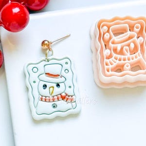 May include: A white snowman earring with a gold post and a matching cookie cutter. The snowman has a black top hat, orange carrot nose, and a red and pink scarf. The earring is rectangular with a scalloped edge.