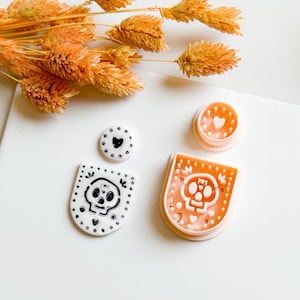 May include: A collection of skull-themed craft supplies. Includes a white and black skull-shaped pendant, a small white circle with a black heart, and a light orange skull-shaped cookie cutter. Dried orange flowers are in the background.