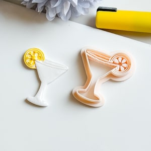 Op de afbeelding: Twee cocktailvormige koekjessnijders. Eén is wit met een gele citroenschijf, de andere is licht perzikkleurig. De uitstekers liggen op een wit oppervlak met een gele pen en een witte bloem op de achtergrond.