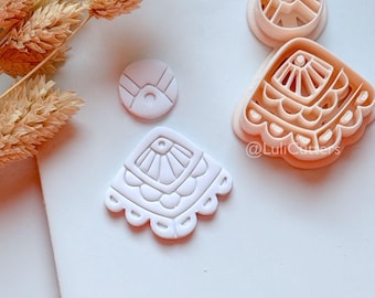 May include: Four light pink cookie cutters in various shapes, including a fan-like design with scalloped edges, a circle, and a geometric shape. The cutters are arranged on a white surface, with dried floral stems in the upper left corner. The text "@LuliCutters" is visible.