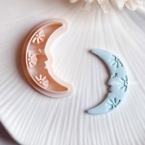 Könnte beinhalten: Ein hellblauer Halbmond-Ausstecher mit floralem Design. Der Ausstecher liegt auf einer weißen Oberfläche neben einem rosa Halbmond-Ausstecher. Der Text '@lulucutters' ist im Bild sichtbar.