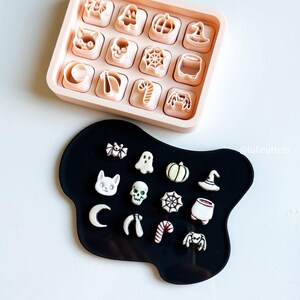 May include: A collection of Halloween-themed cookie cutters in a light pink tray and on a black surface. The cutters include shapes like a cat, pumpkin, spider web, witch hat, and ghost. The cutters are in a variety of shapes and sizes.