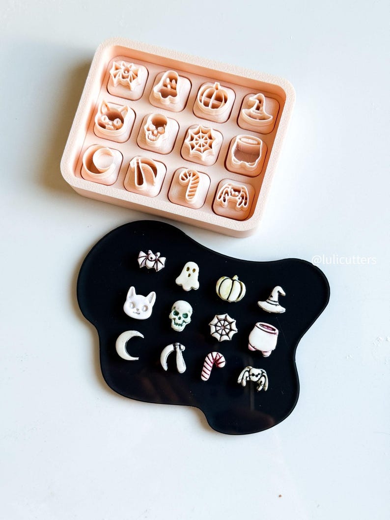 May include: A collection of Halloween-themed cookie cutters in a light pink tray and on a black surface. The cutters include shapes like a cat, pumpkin, spider web, witch hat, and ghost. The cutters are in a variety of shapes and sizes.