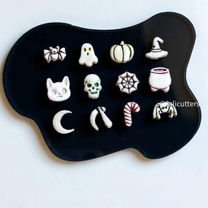 May include: A collection of Halloween-themed charms in white and cream colors, including a bat, ghost, pumpkin, witch's hat, cat, skull, spiderweb, cauldron, crescent moon, scythe, candy cane, and spider, arranged on a black surface.