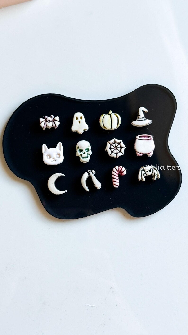 May include: A collection of Halloween-themed charms in white and cream colors, including a bat, ghost, pumpkin, witch's hat, cat, skull, spiderweb, cauldron, crescent moon, scythe, candy cane, and spider, arranged on a black surface.