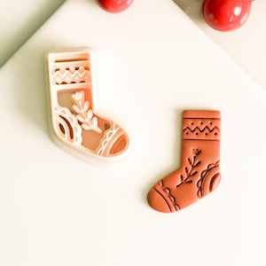 May include: A light brown Christmas stocking cookie cutter and a matching biscuit. The stocking has a floral design and decorative patterns. Red berries are in the background.