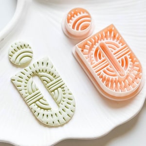 Op de afbeelding: Een verzameling polymeerklei uitsteekvormen en afgewerkte klei oorbellen. De uitsteekvormen zijn perzikkleurig en de oorbellen lichtgroen. De oorbellen en uitsteekvormen hebben een geometrisch ontwerp. De oorbellen liggen op een wit, geschulpte dienblad.