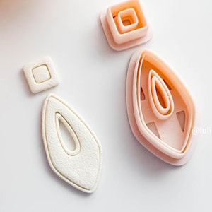 May include: A collection of light pink polymer clay earring cutters in various shapes. The cutters include a square, a square within a square, and an elongated teardrop shape with an oval cutout. The shapes are arranged on a white surface.