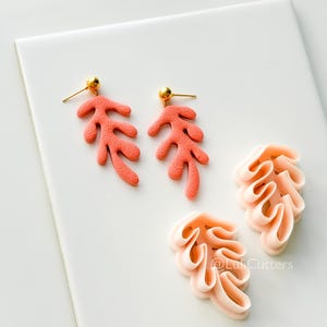 May include: Coral-colored leaf-shaped earrings with gold-colored posts and matching leaf-shaped clay cutters. The earrings and cutters are displayed on a white surface. The cutters have the text "@LukiCutters".