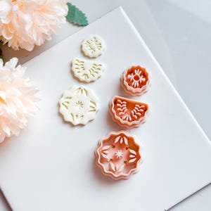 Op de afbeelding: Een verzameling bloemvormige koekjessnijders in wit en perzik. De snijders zijn gerangschikt op een wit oppervlak, met een paar geperste koekjes ernaast. De snijders hebben gedetailleerde bloemmotieven, perfect om te bakken. Perzik kleurige bloemen staan op de achtergrond.