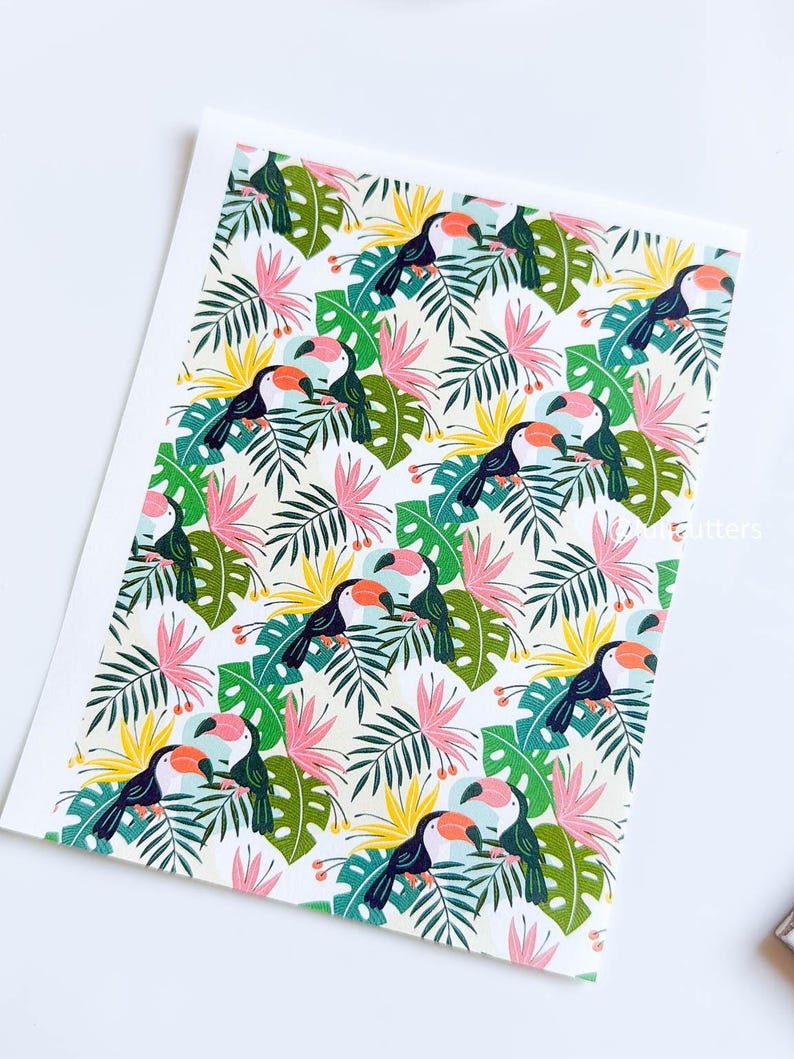 May include: A seamless pattern featuring toucans with orange beaks and black bodies perched on green and yellow tropical leaves. The background is white with pink flowers.