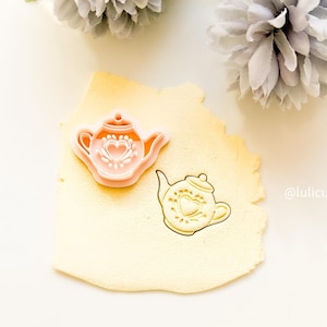 May include: A pink and orange teapot-shaped cookie cutter with a heart design. A gold-colored teapot impression is next to the cutter. The cutter is on a light yellow surface. The image has a soft, bright aesthetic.