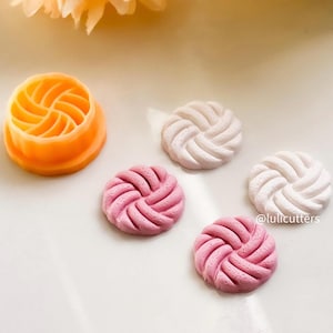 May include: A set of four cookie cutters, two pink, one white and one orange. The cutters are shaped like a braided knot. The orange cutter is a mould for making the cookies.