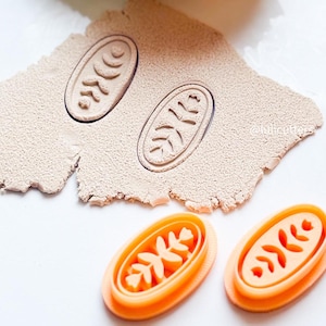 May include: Two orange plastic cookie cutters with a leaf design. The cutters are oval shaped and have a raised design of a leaf with three branches.