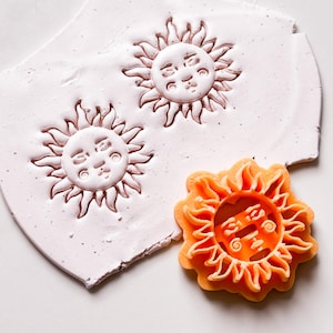 Op de afbeelding: Twee witte zonvormige koekjesvormen met een gezichtsontwerp en één oranje zonvormige koekjesvorm met een gezichtsontwerp.