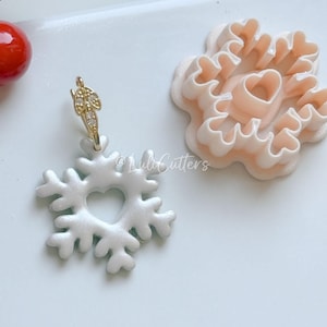 以下が含まれることがあります： ハート型の切り抜きがある白い雪の結晶のイヤリング。ゴールドカラーのイヤリングフックが付いています。隣には、ハート型の中心部がある、薄いピンク色の雪の結晶型のクッキーカッターがあります。白い背景です。