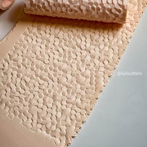 May include: A light beige clay roller with a honeycomb pattern, alongside a sheet of clay with the same embossed design. The roller is cylindrical, and the clay sheet is rectangular. The image is taken from above.