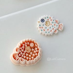 Può includere: Due formine per biscotti a forma di pesce palla. Uno è di colore pesca chiaro, l'altro è bianco con dettagli blu, pesca e oro. Le formine per biscotti hanno disegni dettagliati, tra cui pinne, occhi e punte. Il testo "@LuliCutters" è nell'angolo in basso a destra.