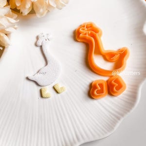 May include: A white goose-shaped fondant with yellow feet and an orange goose-shaped cookie cutter. The cookie cutter and fondant are on a white plate. The text "@lulicutters" is visible.