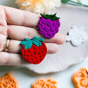 May include: Two silicone molds for baking, one shaped like a strawberry and the other like a raspberry. The strawberry mold is red with green leaves, and the raspberry mold is purple.