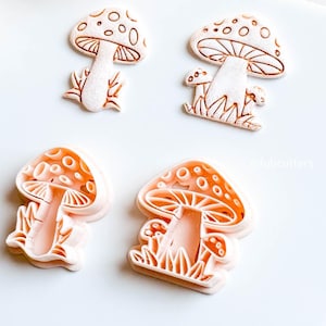 May include: Four mushroom-shaped cookie cutters. Two are white with brown outlines, and two are orange with white outlines. The cutters have detailed designs of mushroom caps, stems, and grass. The image is on a white surface.