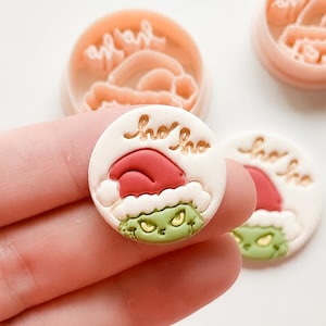 Puede incluir: Una pieza circular de arcilla blanca con un personaje verde de ojos amarillos que lleva un gorro de Papá Noel rojo, con el texto "ho ho" en relieve. Dos cortadores de plástico redondos de color melocotón se ven al fondo.