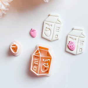 May include: A selection of strawberry-themed cookie cutters and stamps in white and orange. The designs feature milk cartons and strawberries. The items are arranged on a white surface, with some artificial flowers in the background.