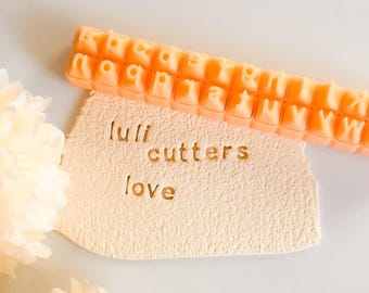 May include: An orange alphabet stamp tool is positioned above a piece of beige clay with the words "luli cutters love" imprinted. The stamp tool is filled with the letters of the alphabet. The clay has a textured surface.