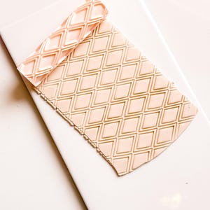 May include: A light peach-coloured clay slab with a diamond pattern, alongside a matching rolling pin. The rolling pin is embossed with the same diamond design. The items are on a white surface.