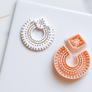 May include: Two earrings, one white and one peach, with geometric designs. The white earring is a hoop with gold accents and a square top. The peach earring is a cutter with a similar design. Both have a tribal or bohemian aesthetic.