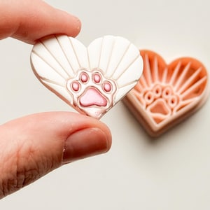 May include: Two heart-shaped decorative pieces. One is held by a person's fingers. The held piece is white with a pink paw print design. The other piece is peach-colored with a paw print design.