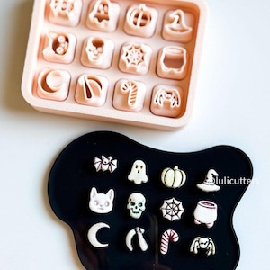 Op de afbeelding: Een lichtroze dienblad met 12 Halloween-thema koekjessnijders, waaronder een spook, pompoen en heksenhoed. Een zwart, onregelmatig gevormd dienblad bevat 12 versierde Halloween-koekjes, waaronder een spin, schedel en zuurstok.