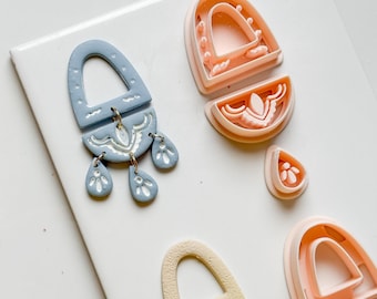 Könnte beinhalten: Eine Sammlung von Polymer-Ton-Ohrringkomponenten und -ausstechern. Die Ohrringe sind in verschiedenen Stadien der Fertigstellung, mit einem hellblauen Ohrring mit weißen floralen Details und goldfarbenen Sprungringen. Weitere Komponenten sind pfirsich- und cremefarben.