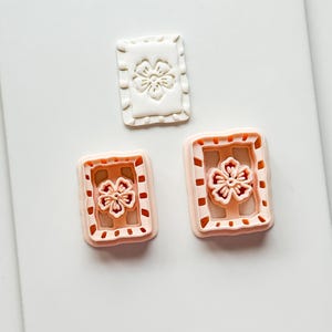 May include: Three rectangular cookie cutters. One is white, with a floral design in the center. The other two are peach-colored, with a floral design and a dotted border. These cutters are likely for baking or crafting.