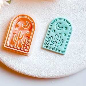 May include: Two cookie cutters with a desert theme. One is orange, the other is teal. Each has a cactus, crescent moon, and stars design within an arched frame. The cutters are made of plastic and sit on a white surface.