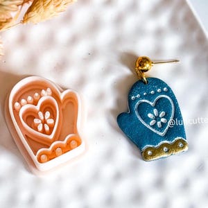 Peut inclure: Boucle d'oreille en forme de moufle bleue avec des détails blancs et dorés, ornée d'un cœur et d'une fleur. À côté, un emporte-pièce en forme de moufle pêche, avec le même motif.