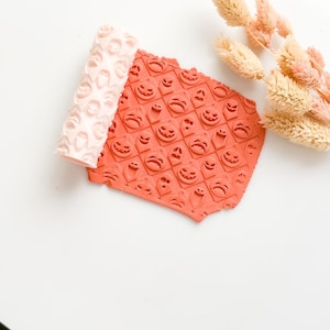 Op de afbeelding: Een lichtroze deegroller en een stuk oranje klei met een herhalend patroon van pompoengezichten. De deegroller heeft een vergelijkbaar patroon. Gedroogde bloemen op de achtergrond. Halloween-thema knutselbenodigdheden.