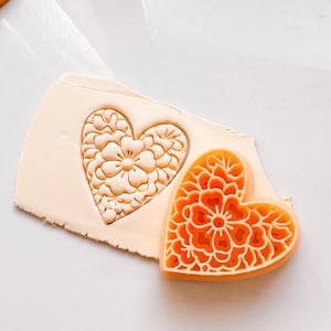 Op de afbeelding: Een close-up van een oranje hartvormige mal met een gedetailleerd bloemenpatroon. De mal wordt in een lichtbeige kleioppervlak gedrukt, waardoor een afdruk van het patroon ontstaat.