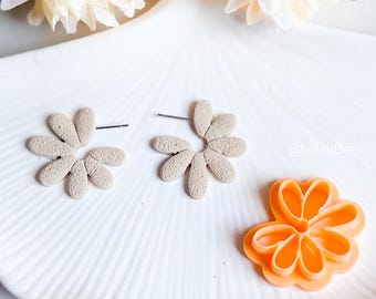 Peut inclure: Deux boucles d'oreilles en forme de fleur beige avec une surface texturée, et un petit emporte-pièce en forme de fleur orange sur une assiette blanche. Le texte '@lulucutters' est visible en arrière-plan.