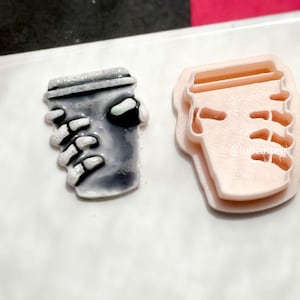 May include: Two coffee-themed cookie cutters. One is a grey coffee cup held by a skeleton hand. The other is a light pink outline of a coffee cup with a hand holding it. The background is white.