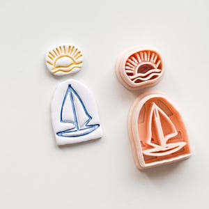 Op de afbeelding: Een verzameling koekjessnijders met nautisch thema. Bevat een witte en blauwe zeilboot, een gele zon met golven en twee roze koekjessnijders met een zeilboot- en zonontwerp. Perfect voor zomerse bakkerij.