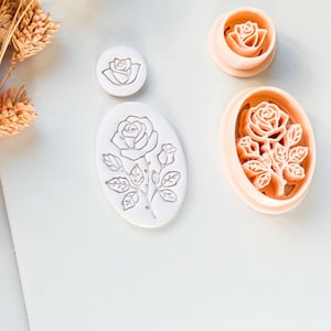 Könnte beinhalten: Eine Sammlung von Ausstechern und Stempeln in Rosenform. Die Ausstecher sind pfirsichfarben und die Stempel sind weiß. Die Designs umfassen eine kleine Rose, eine Rose in einem Kreis und eine Rose mit Stiel und Blättern.